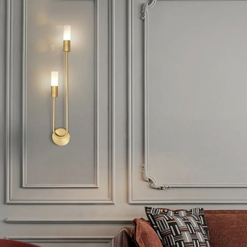 A contemporary Nordic stick on wall light by XCEPTION, featuring a gold finish and two frosted glass light covers, is mounted on a grey paneled wall. In the foreground, a brown sofa adorned with patterned pillows contributes warmth to the space.