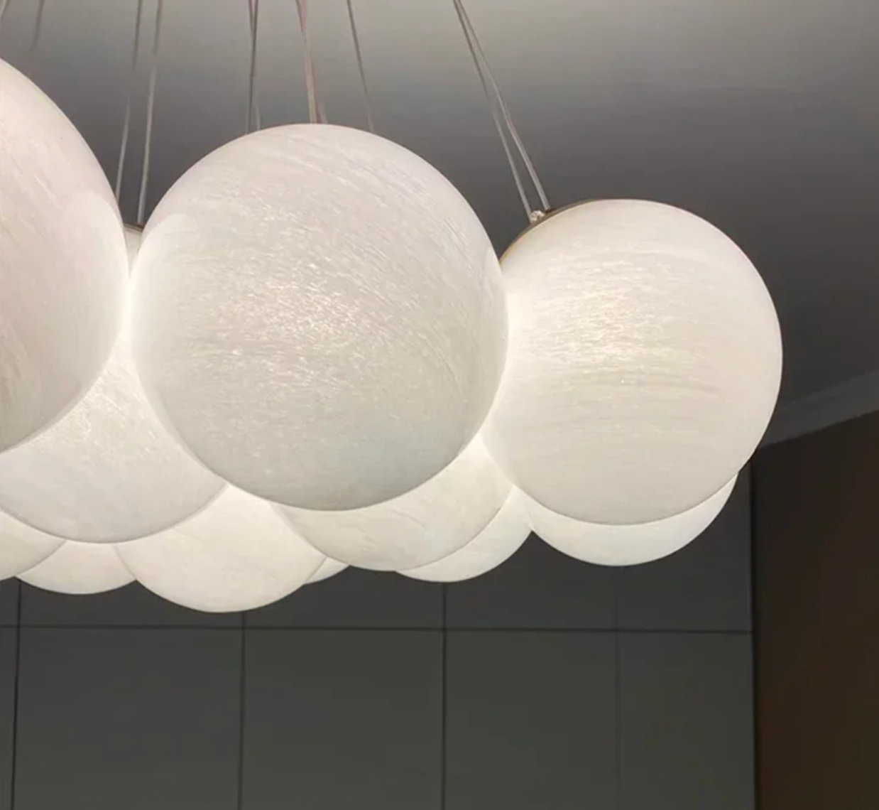 A modern chandelier, known as the Blurred Bubbles Chandelier by XCEPTION, featuring a series of spherical, white, glowing lamps hangs elegantly from the ceiling. Complementing this design is a neutral-colored wall and ceiling in the background, which enhances the minimalist aesthetic of the space.