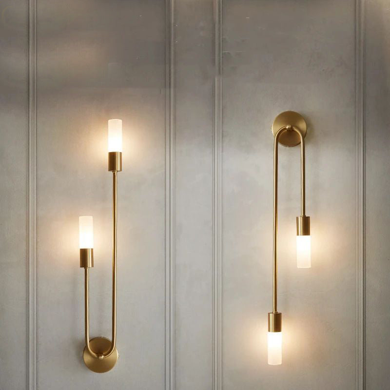 A pair of Nordic stick on wall lights from XCEPTION enhances a light-colored panel wall. These modern sconces incorporate energy-saving LED technology, showcasing two cylindrical lights linked by elegant, curved gold-finished metal arms, providing a warm glow.
