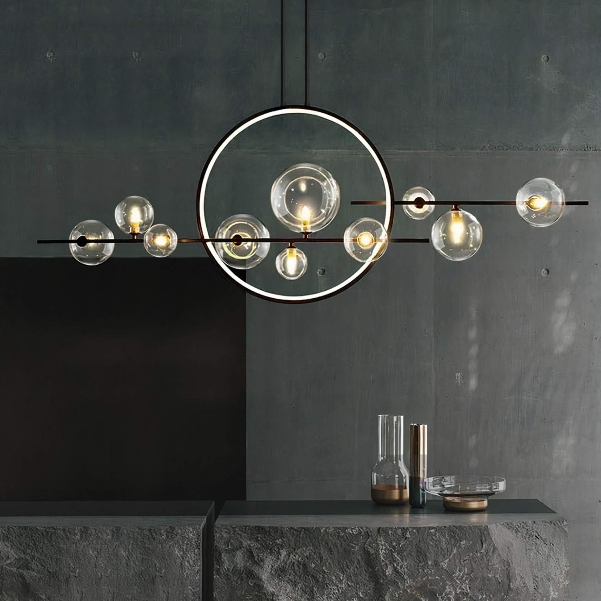 The Comos Ceiling Light by XCEPTION, featuring circular and spherical glass elements, hangs from a ceiling against a dark green-gray wall. Below, a minimalist table displays two glass vases, one tall and the other short, accentuating the contemporary aesthetic.