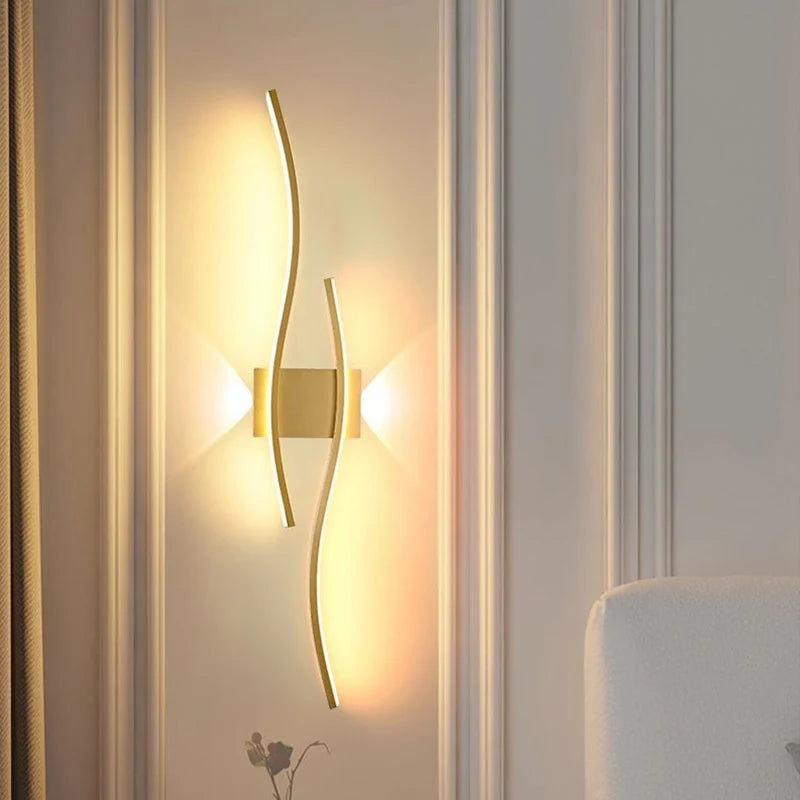 The Minimalisto Wall Light by XCEPTION is a modern wall lamp that features two sleek gold strips and minimalist LED lights, casting a warm glow on a textured wall. Its design creates a soft ambiance around the fixture, enhancing the room's elegance. A portion of a white sofa can be seen in the corner.