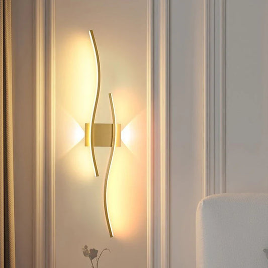 The Minimalisto Wall Light by XCEPTION is a modern wall lamp that features two sleek gold strips and minimalist LED lights, casting a warm glow on a textured wall. Its design creates a soft ambiance around the fixture, enhancing the room's elegance. A portion of a white sofa can be seen in the corner.