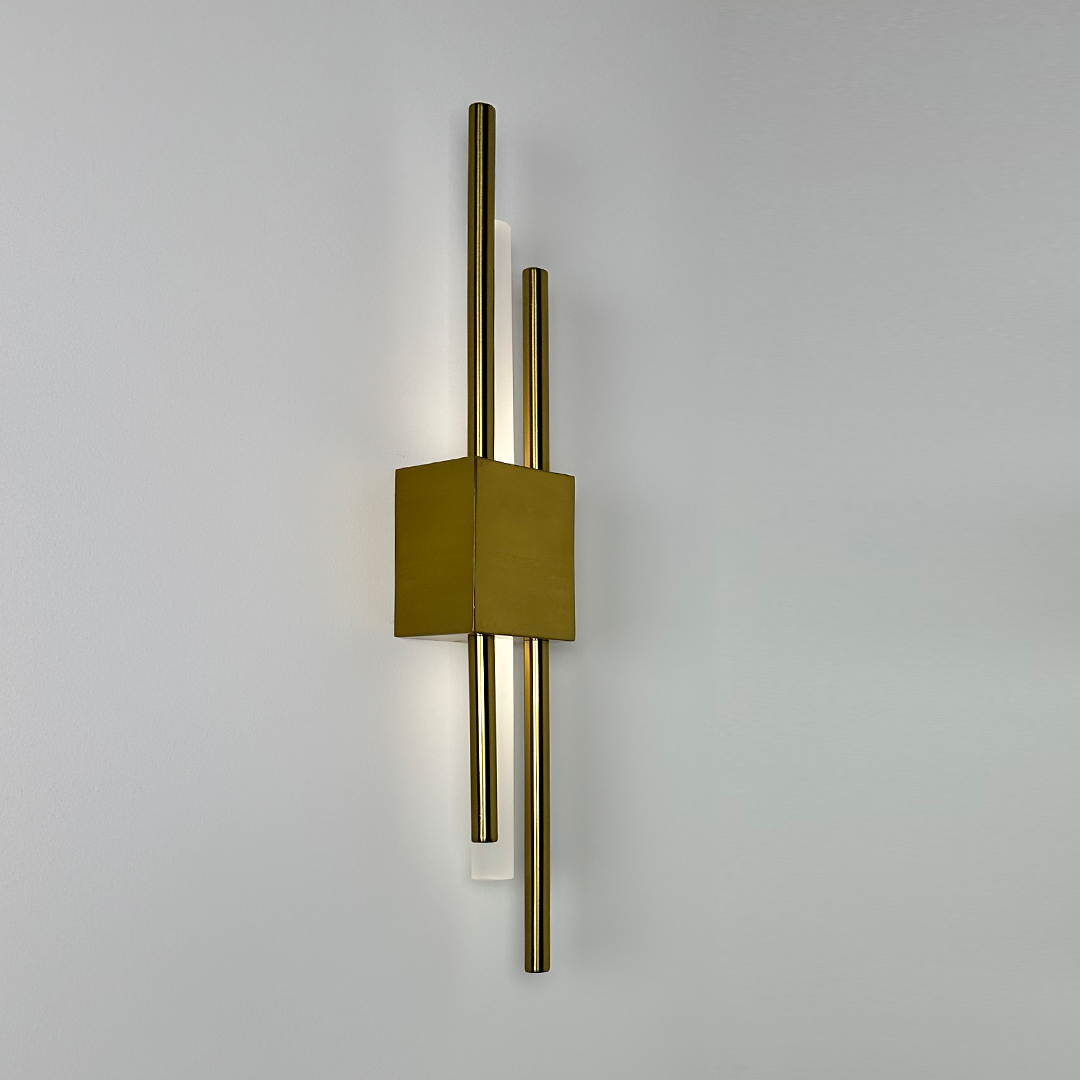 The Glitch Wall Light by XCEPTION, in a elegant gold finish, showcases a minimalist design with two parallel brass rods extending vertically through a sleek rectangular block. This wall-wired fixture emits soft light that enhances a plain white wall with a warm and sophisticated ambiance.