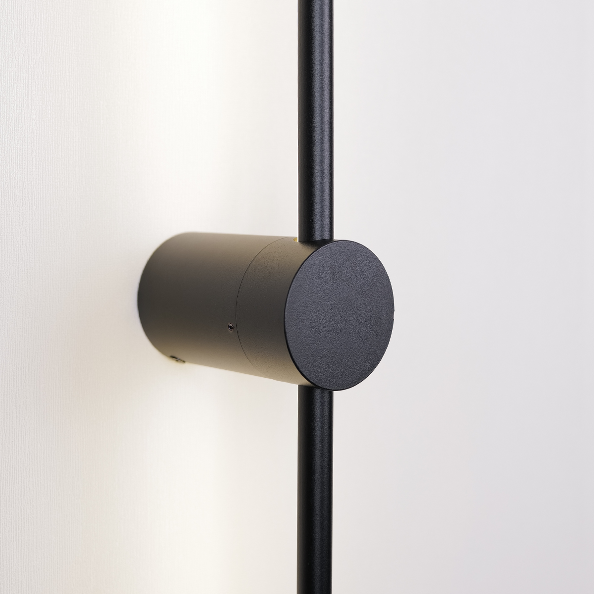 XCEPTION's Nordic Light Stick • 2-Pack features a minimalist black wall-mounted lamp with a cylindrical base and vertical rod, elegantly casting soft light on a white wall while exuding a modern design aesthetic.