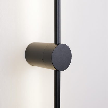 XCEPTION's Nordic Light Stick • 2-Pack features a minimalist black wall-mounted lamp with a cylindrical base and vertical rod, elegantly casting soft light on a white wall while exuding a modern design aesthetic.