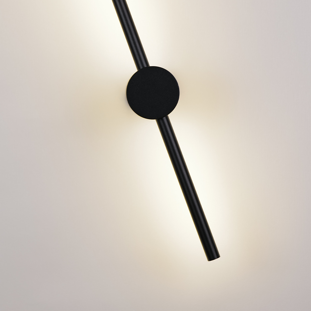 The Nordic Light Stick • 2-Pack from XCEPTION showcases a contemporary wall lamp design with a minimalist black rod that elegantly intersects a circular black base, casting a gentle, warm glow against a plain white wall.