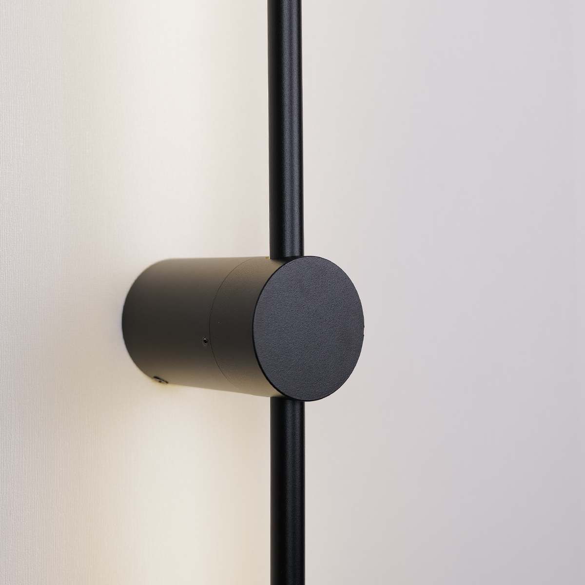 Close-up of a Nordic Light Stick by XCEPTION, a modern wall-mounted black cylindrical light fixture on a white wall. This fixture features a sleek vertical rod with a round, cylindrical base that casts soft light onto the surface.