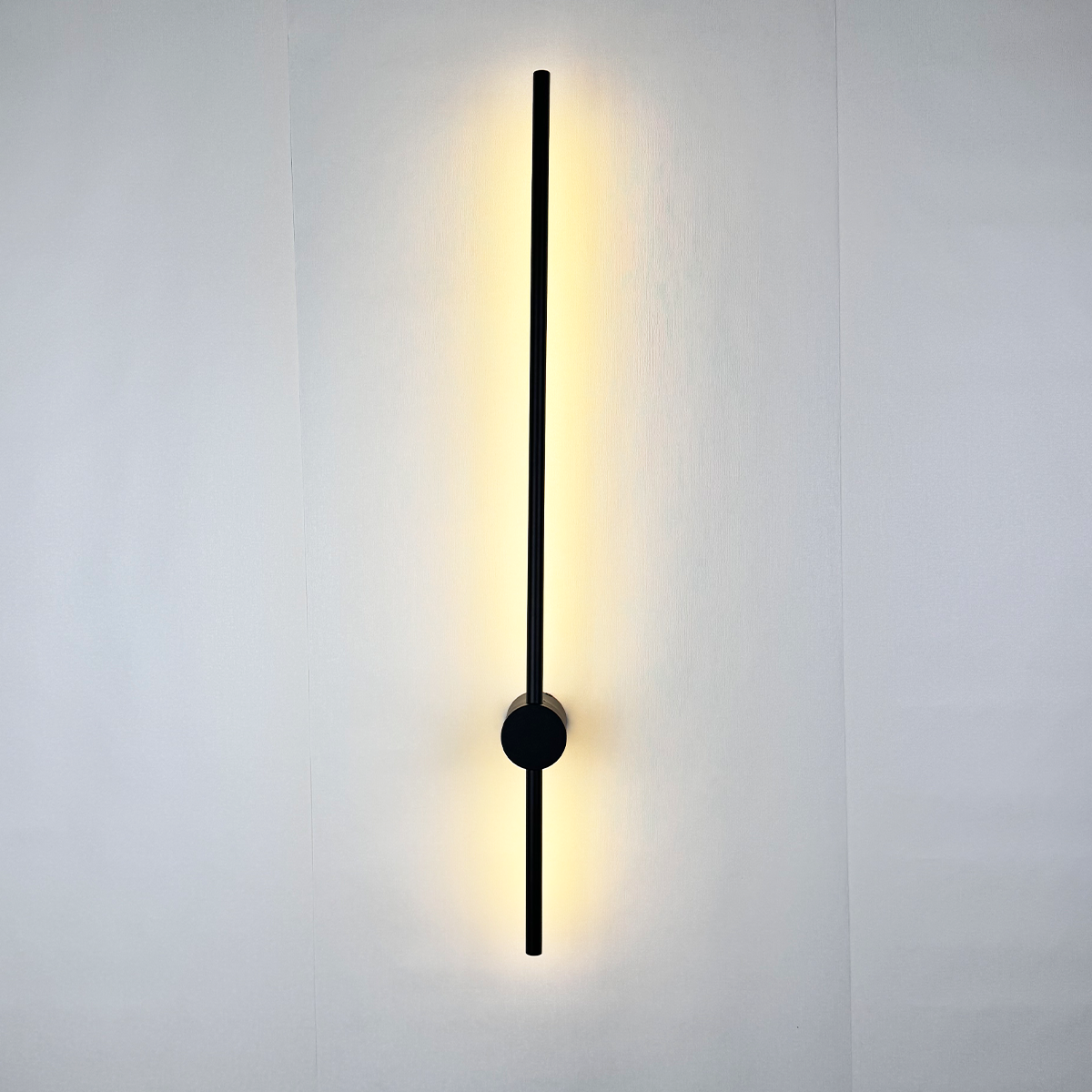 The "Nordic Light Stick • 2-Pack" by XCEPTION features a sleek, contemporary wall lamp with a vertical black rod design, ideal for mounting on a white surface. It casts a gentle, warm yellow light that bathes the surrounding area in a subtle glow.