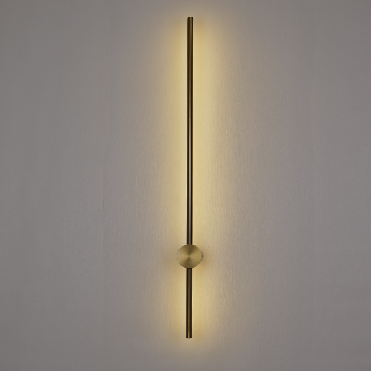 The Nordic Light Stick • 2-Pack by XCEPTION features a sleek, vertical wall light fixture with a long, slender metallic rod that casts a warm glow on a plain backdrop. A small sphere intersects the rod at the center, contributing to its simple yet sophisticated design.