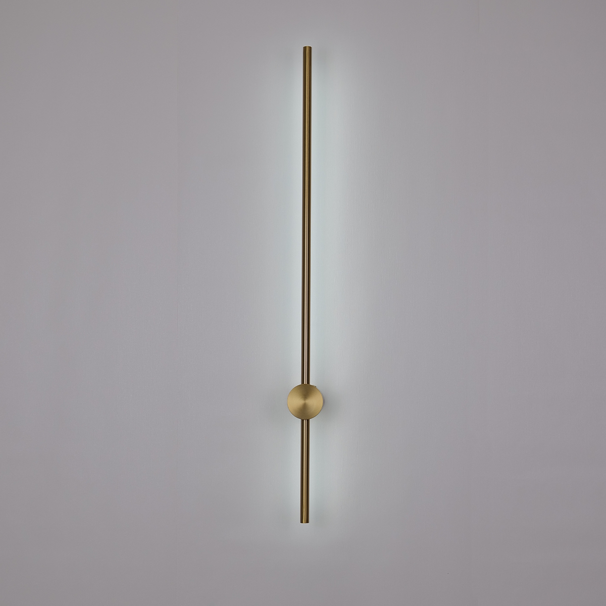 The Nordic Light Stick • 2-Pack, by XCEPTION, showcases a modern, minimalist design featuring a vertical brass rod and a central round element. It emits a soft light from behind, casting a subtle glow on the pale wall.