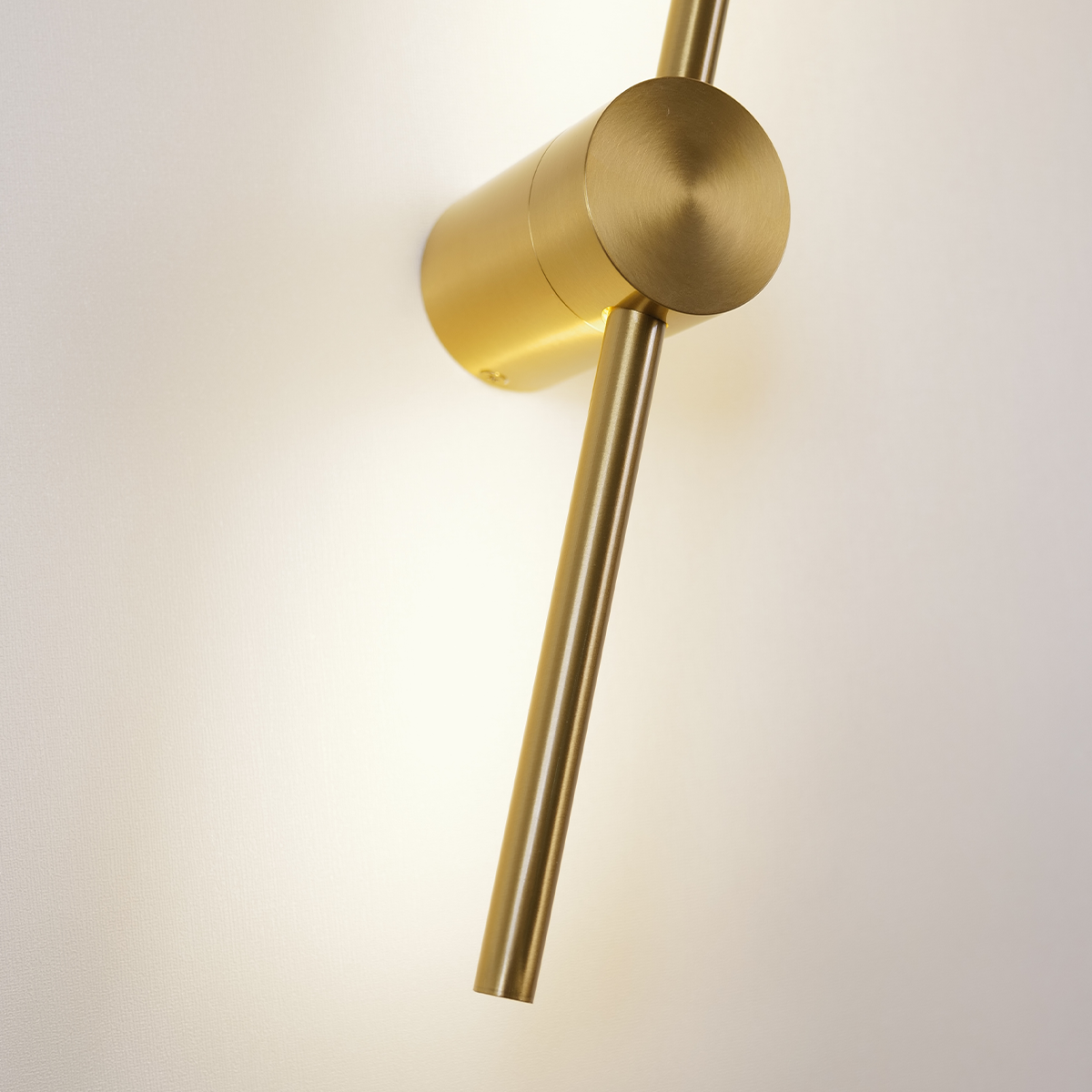 Close-up of XCEPTION's Nordic Light Stick from the 2-Pack collection, featuring a modern gold wall sconce with a cylindrical base and an elongated arm extending diagonally. The light casts a warm glow on the white wall, highlighting the fixture's sleek, minimalist design.