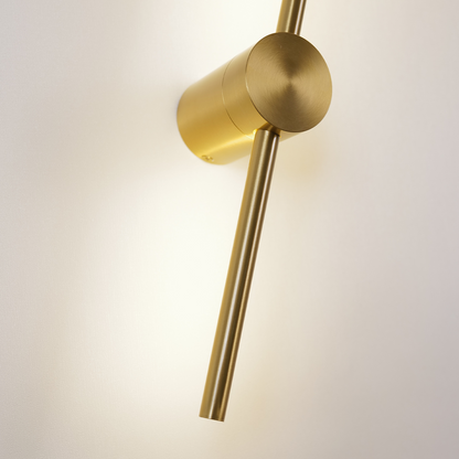 Close-up of XCEPTION's Nordic Light Stick from the 2-Pack collection, featuring a modern gold wall sconce with a cylindrical base and an elongated arm extending diagonally. The light casts a warm glow on the white wall, highlighting the fixture's sleek, minimalist design.