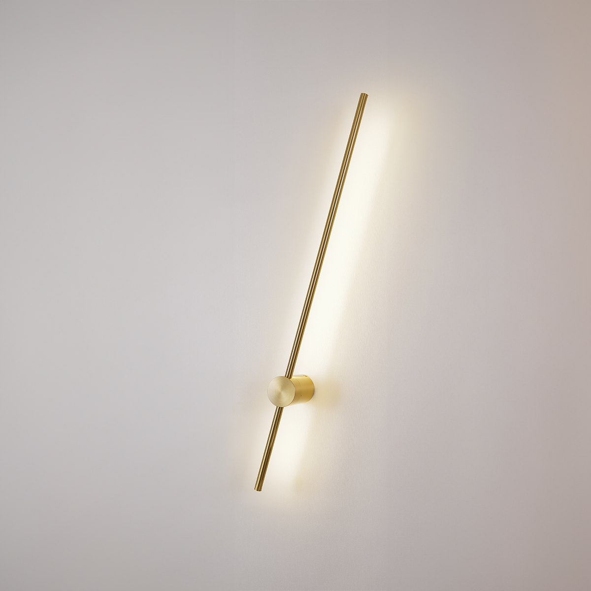 The Nordic Light Stick • 2-Pack by XCEPTION offers a modern design featuring a slim, vertical gold rod that emits a warm and soft light against a plain white wall. The minimalistic style includes a cylindrical mount holding the rod at a slight angle.