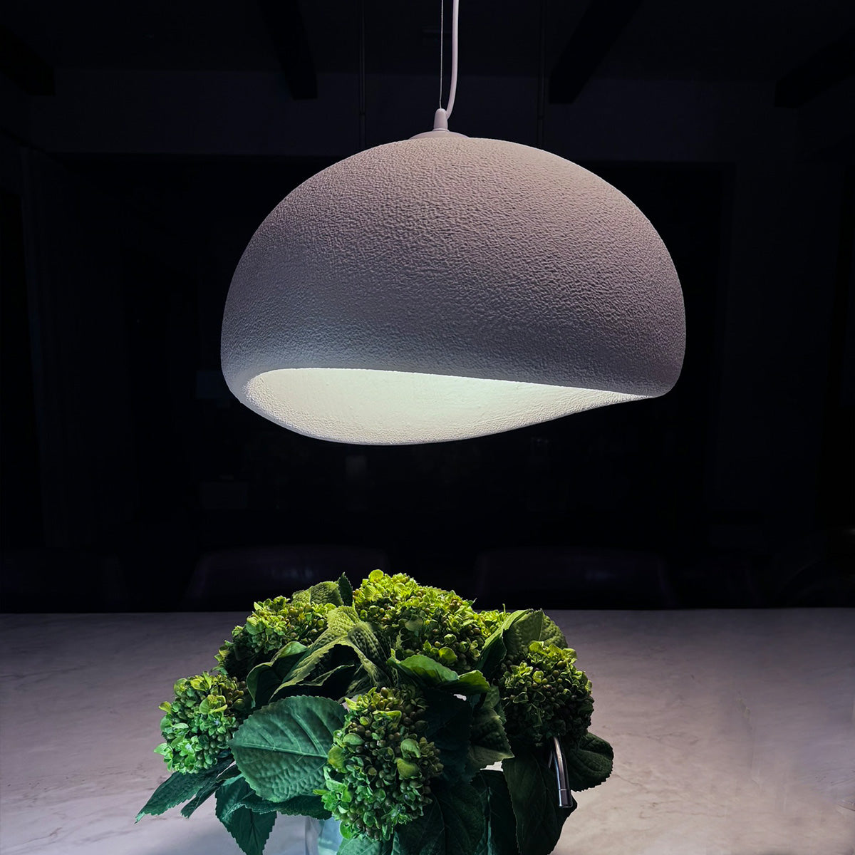 The Organic Cove by XCEPTION, a contemporary pendant lamp featuring a textured, dome-shaped shade, casts a warm glow over a dining table. Below, a vibrant bouquet of green hydrangeas enhances the cozy and elegant ambiance.