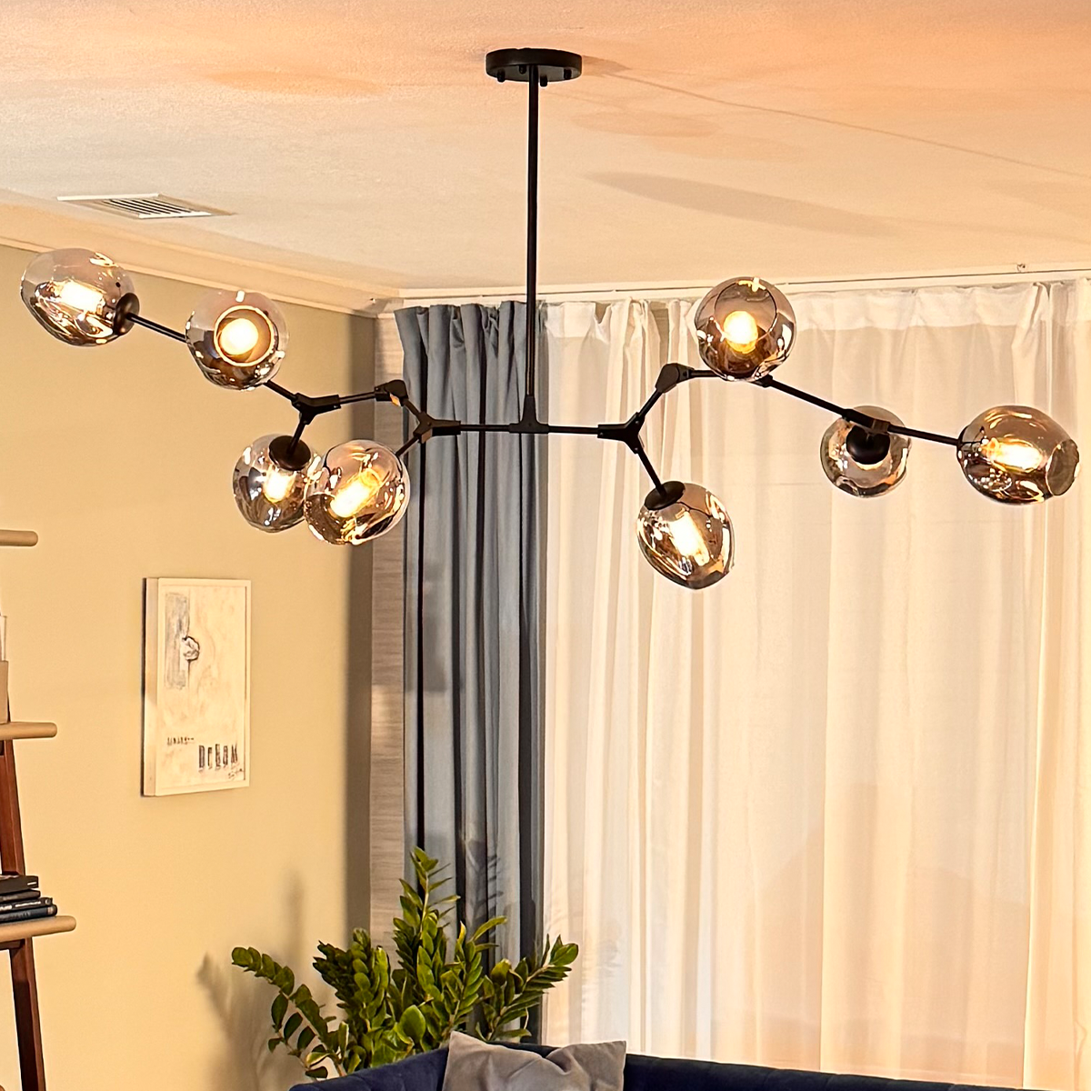 A Retro Loft Ceiling Chandelier in black from XCEPTION, featuring eight globe-shaped reflective bulbs, hangs from the ceiling in a living room. The room includes beige walls, a blue sofa, a leafy plant, and a white and blue curtain.