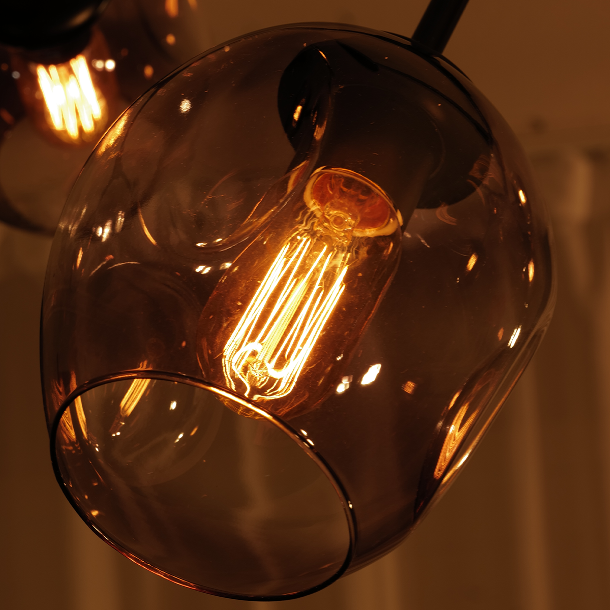 Introducing the Retro Loft Ceiling Chandelier in Black by XCEPTION, featuring a glowing incandescent light bulb enclosed in a rounded, amber-tinted glass fixture. This piece emits a warm light that enhances the texture and color of the glass, creating a cozy atmosphere.