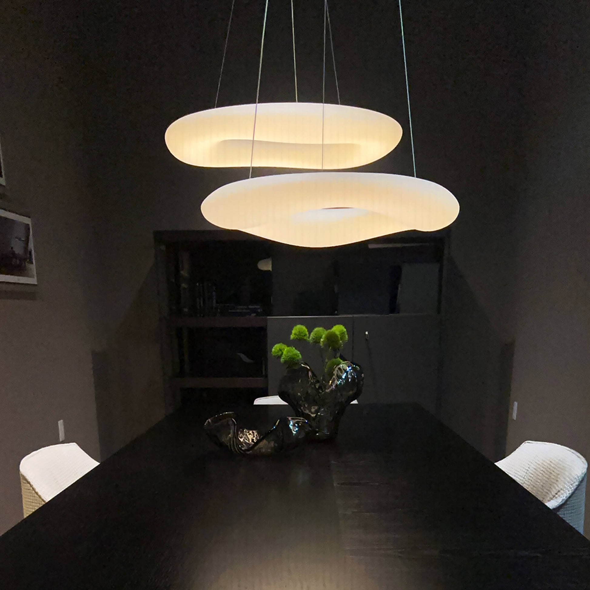 A contemporary dining room features a dark wooden table accompanied by two white chairs. A sculptural centerpiece with green plants is elegantly positioned in the center. Overhead, the XCEPTION Cloud Ceiling Light casts a warm glow, enhancing the sleek and minimalist decor with its unique layered design.