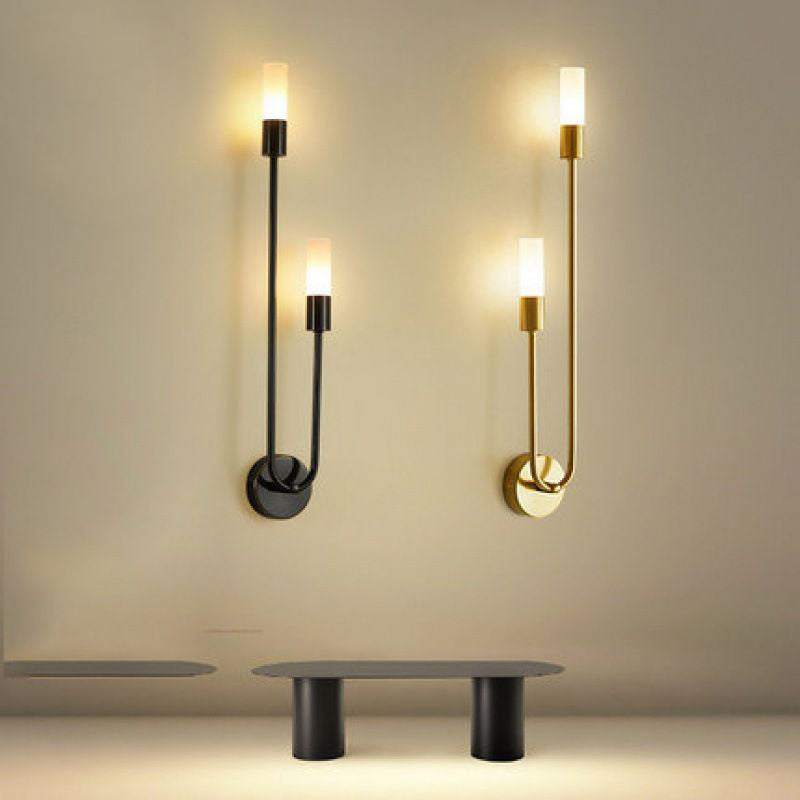 In a minimalist room, two XCEPTION Nordic stick on wall lights, one in black and the other in gold, cast a gentle, warm glow with their energy-efficient LED technology. Below these is a sleek modern bench in black with a slim top and cylindrical legs, all positioned against a beige wall.