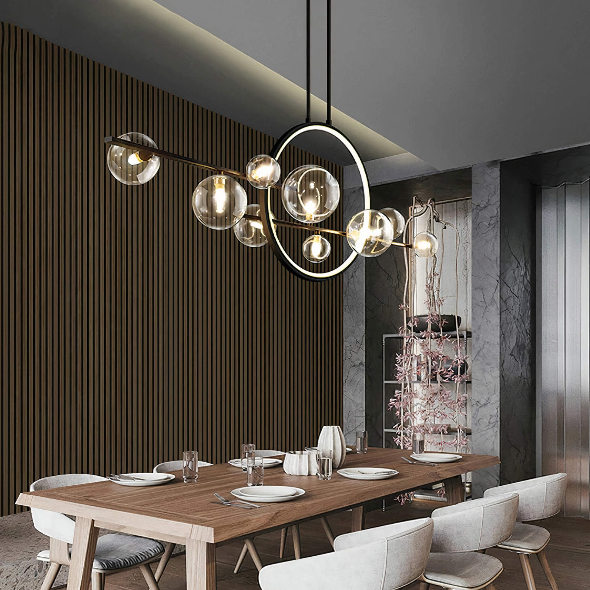 A modern dining room features a wooden table set for six, with the Comos Ceiling Light by XCEPTION hanging above. This artistic chandelier boasts circular frames and spherical lights. The walls combine textured wood and marble, complemented by a decorative plant feature in the background.