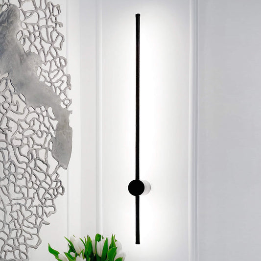 The Nordic Light Stick • 2-Pack by XCEPTION features a stylish design with a vertical black rod and circular base, casting a gentle glow. Nearby, abstract art adorns the wall, while green plants peek into view at the bottom.