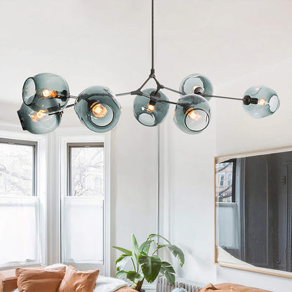 A contemporary living room showcasing the XCEPTION Retro Loft Ceiling Chandelier • Black, distinguished by its multiple glass globes suspended from a black metal frame. The space is complemented by large windows, a comfortable sofa adorned with cushions, and a green plant in the corner.