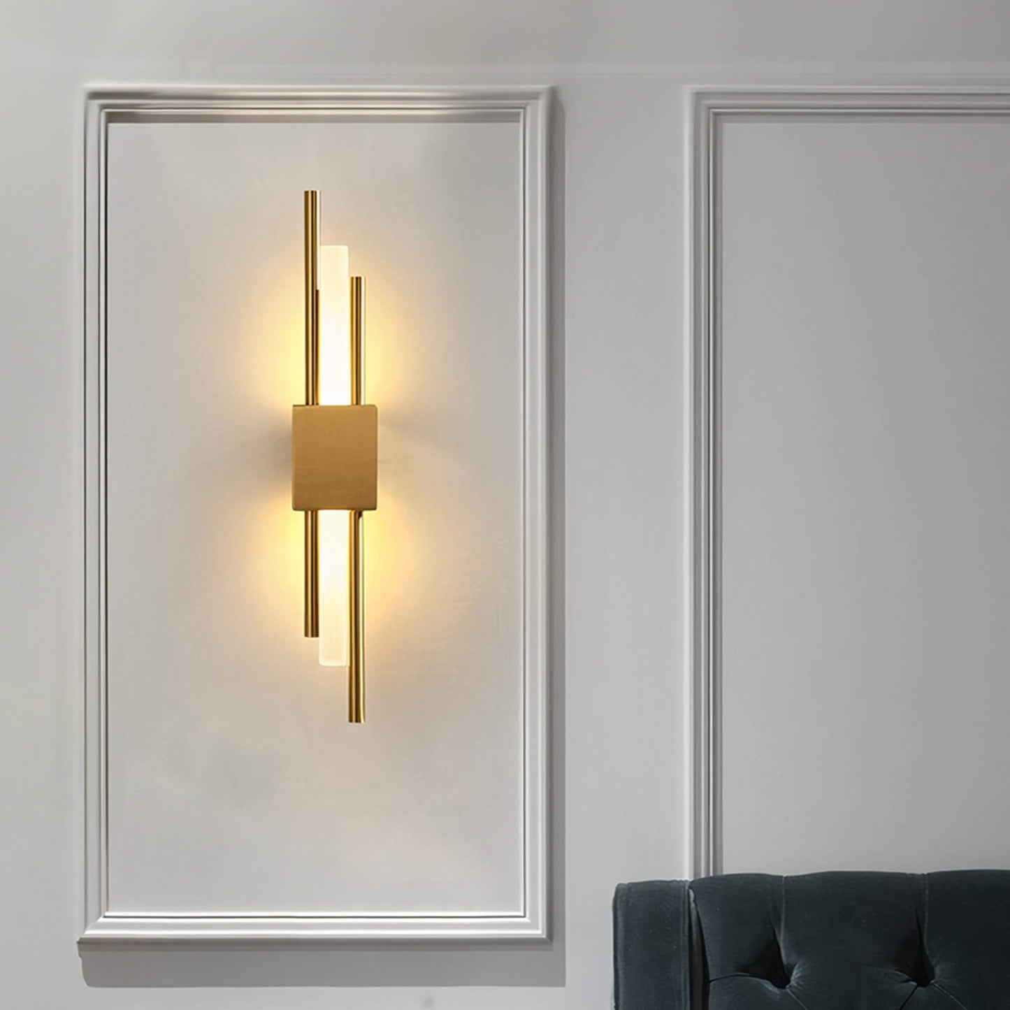 A minimalist wall featuring the Glitch Wall Light by XCEPTION, a geometric gold-toned fixture that casts a warm glow with its contemporary design of vertical lines. Beside it, the edge of a dark green upholstered chair is partially visible.