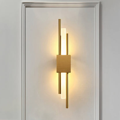 The Glitch Wall Light by XCEPTION features a minimalist design with vertical gold rods and a rectangular central fixture that emits warm light, all beautifully mounted on a white wall adorned with a decorative frame.