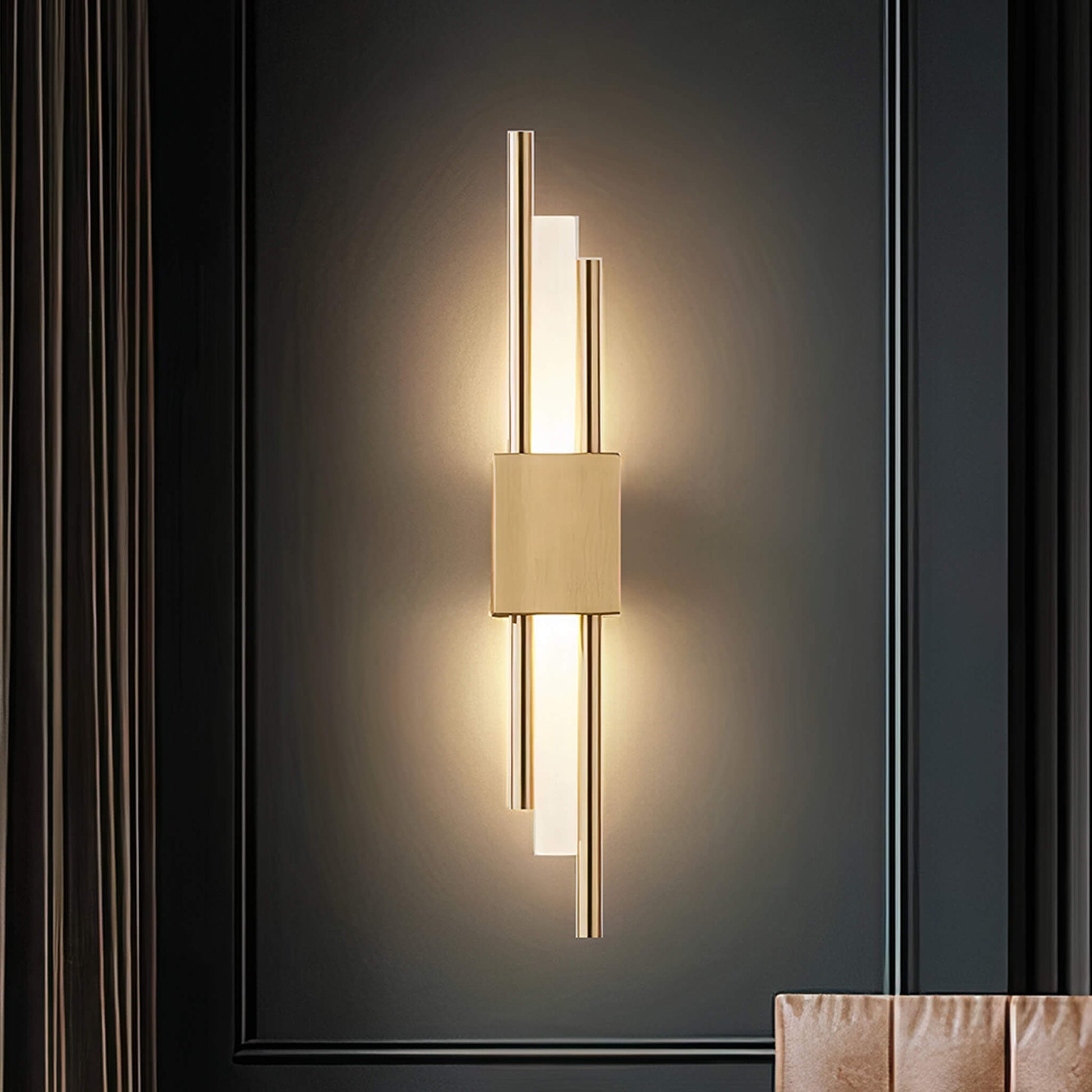The Glitch Wall Light • Gold • Wall Wired by XCEPTION emits a warm glow in a dark room. This modern fixture features a rectangular metallic body with vertical rods extending above and below, casting soft illumination on the wall. A partial view of a chair can be seen at the bottom right.