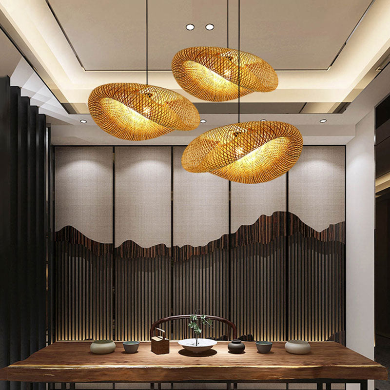 A contemporary dining room showcases a wooden table encircled by chairs. Three oval-shaped pendant lights from the XCEPTION Living Room Ceiling Lights collection, crafted from bamboo, hang overhead, emitting a cozy glow. The wall is adorned with a textured design reminiscent of mountains, enhancing the subtle Southeast Asian style ambiance.
