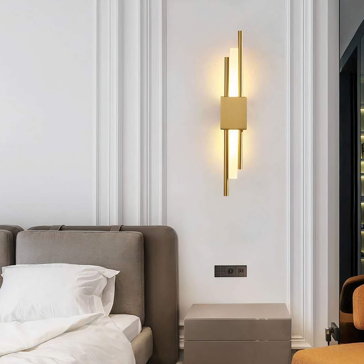 A contemporary bedroom features a beige upholstered bed accompanied by crisp white bedding and the Glitch Wall Light in gold by XCEPTION, adding a touch of geometric elegance to the space. Beside the bed, a small square side table complements the decor, while an orange chair peeks into view on the right. The walls maintain a simple and elegant appearance.
