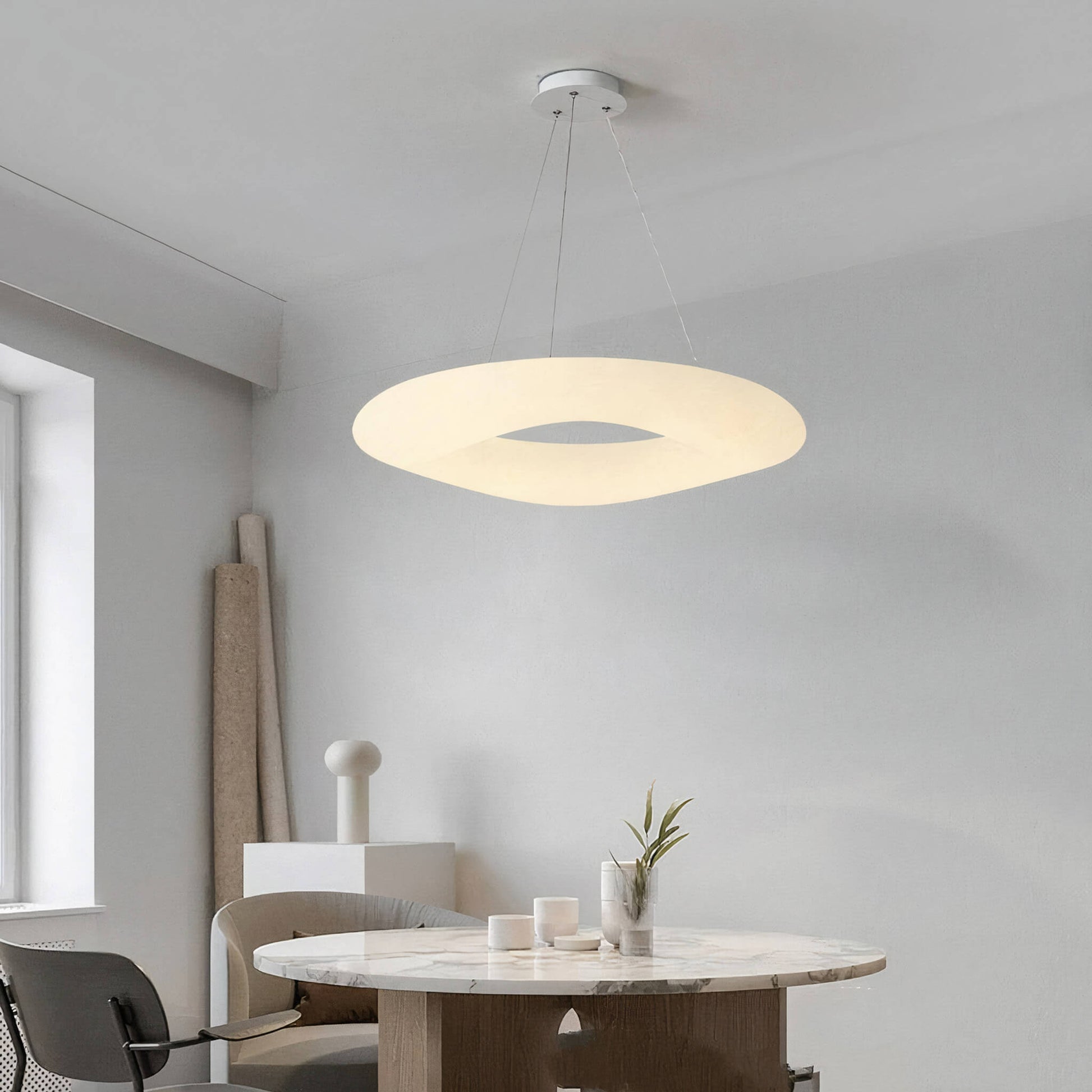 A modern dining area features a round marble table with a wooden pedestal base, complemented by two chairs. Above, the XCEPTION Cloud Ceiling Light casts a circular, halo-like glow. The minimalist decor includes candles and a small plant, while soft natural light pours in through the window.
