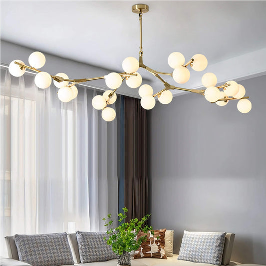 A modern living room features the XCEPTION Heritage Tree Branch Chandelier in gold, showcasing multiple glowing spherical lights. The room is adorned with gray walls, a large window dressed in sheer curtains, a sectional sofa accented with patterned pillows, and a plant placed on the coffee table.