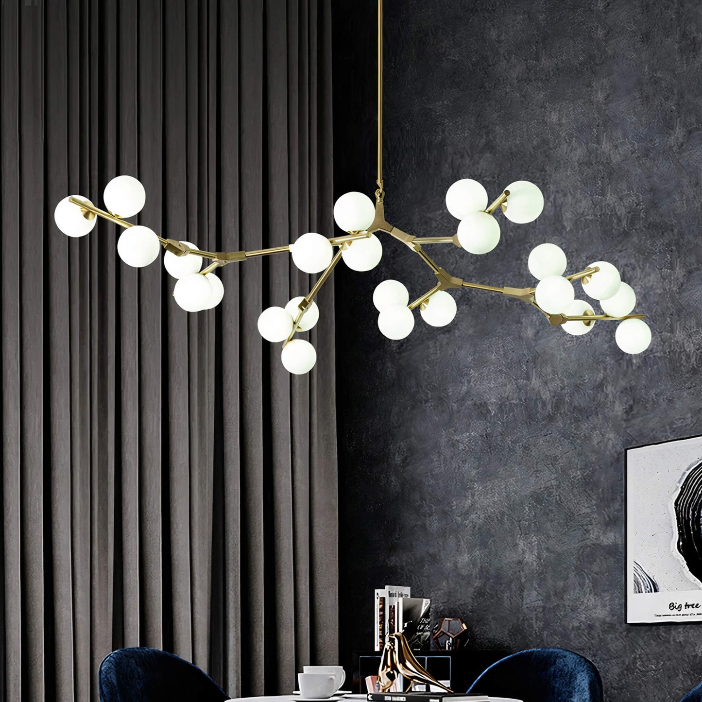 A Heritage Tree Branch Chandelier in gold from XCEPTION, featuring multiple spherical bulbs suspended from its branch-like frame, is elegantly displayed against a dark curtained backdrop with a textured wall and a black-and-white framed artwork. A hint of a blue velvet chair can be seen nearby.