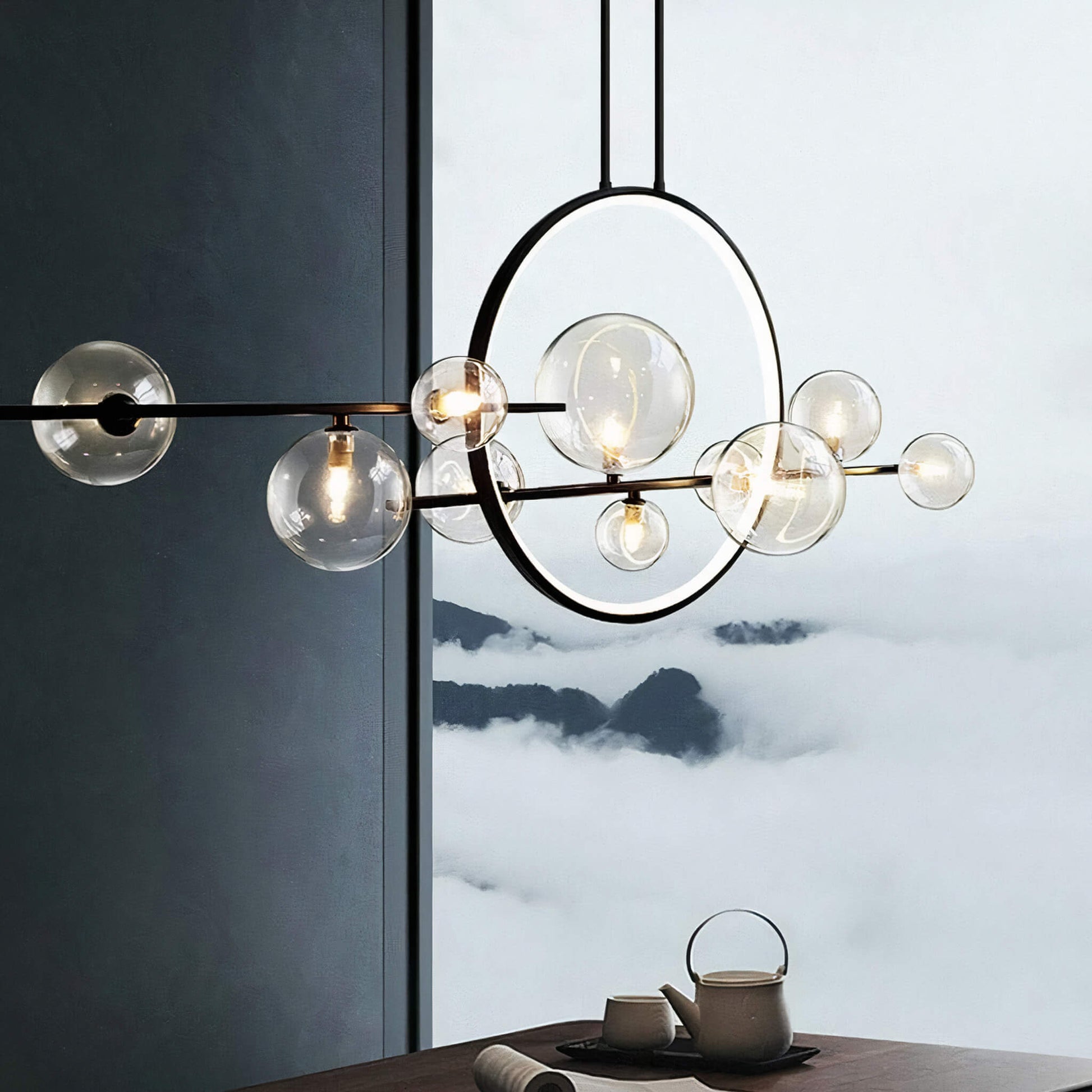 Hanging in front of a large window is the Comos Ceiling Light by XCEPTION, featuring spherical glass bulbs and a circular metal frame. Outside, you can see fog-covered mountains, while below, a tea set rests on a wooden table.