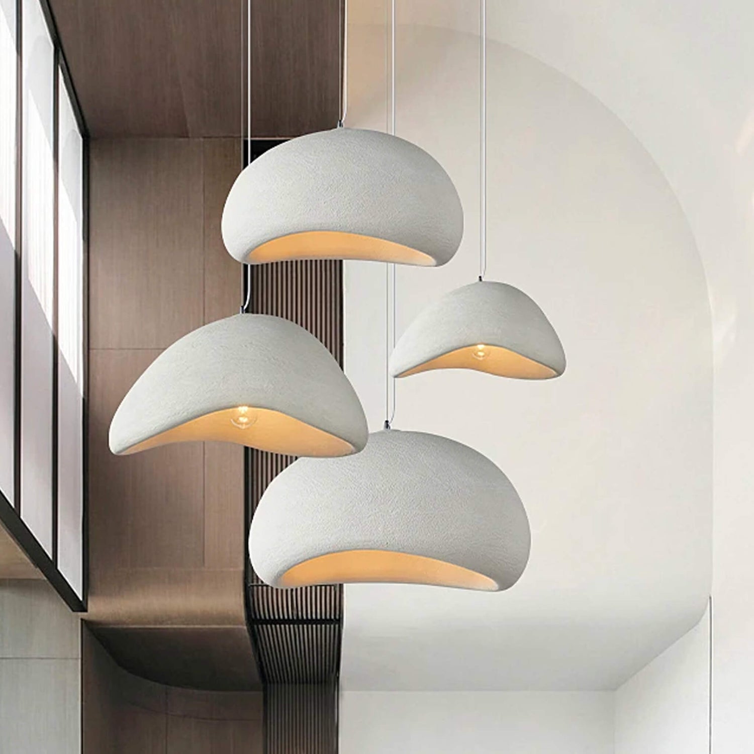 Four modern, dome-shaped pendant lights from XCEPTION's Organic Cove collection hang at varying heights in a minimalist interior. The gray fixtures emit a warm inner glow, beautifully contrasting with the beige wall and large mirror, certainly enhancing the space's serene ambiance.