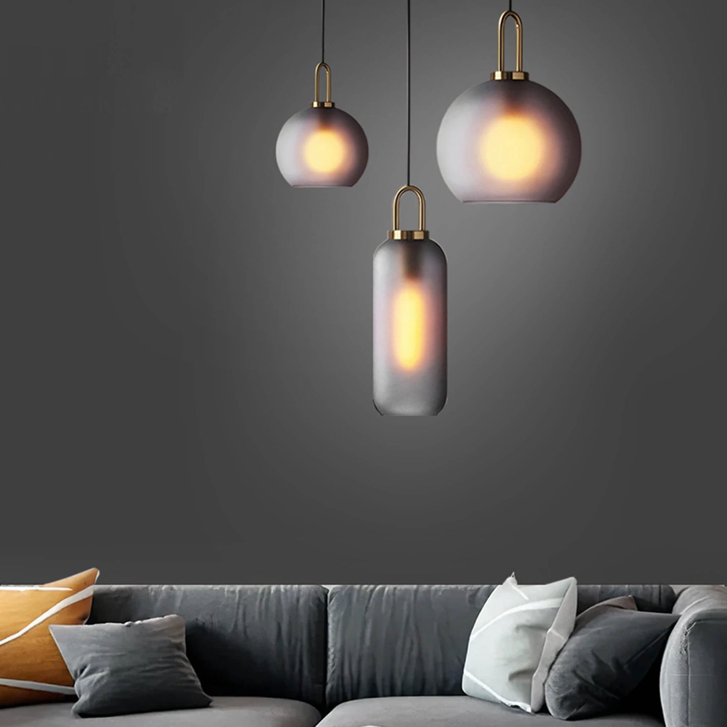 A modern living room featuring a dark gray sofa adorned with assorted pillows is beautifully lit by three XCEPTION Smoked Glass Pendant Light Loft Ceiling fixtures, their frosted glass shades glowing warmly against the dark gray wall to create a cozy atmosphere.