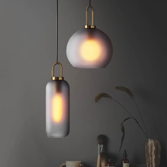 Two Smoked Glass Pendant Light Loft Ceiling fixtures by XCEPTION hang against a gray wall. One features a spherical design, while the other is elongated. A vase filled with dried plants and several cosmetics rest on a table beneath them. The lights cast a warm glow, creating a cozy atmosphere.
